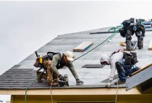 A roofer in a red shirt repairs a shingle roof, with exposed underlayment and stacks of pink roofing materials nearby.