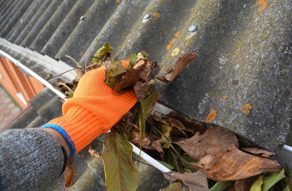 A gloved hand removes leaves and debris from a clogged roof gutter beside weathered roofing tiles.