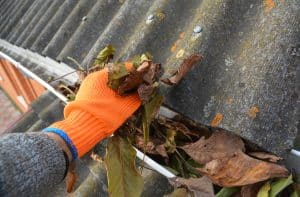 A gloved hand removes leaves and debris from a clogged roof gutter beside weathered roofing tiles.